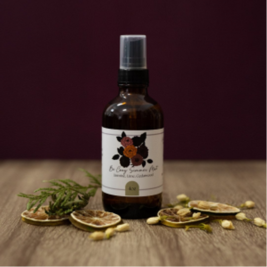 A bottle of Be Cozy Simmer Mist fragrance spray on a wooden surface with citrus slices and cedar.